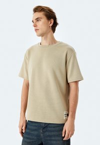 T-shirt leggera in cotone beige con maniche corte, scollatura rotonda e un'etichetta del marchio visibile sull'orlo, indossata con jeans blu scuro.