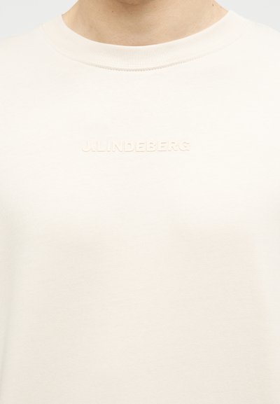 Κοντινό ενός ατόμου που φοράει ανοιχτό μπεζ μπλουζάκι με στρογγυλή λαιμόκοψη και ανάγλυφο κείμενο "J.LINDEBERG" στο στήθος.