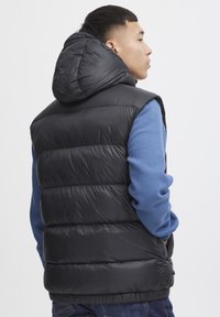 Schwarze Pufferweste mit Kapuze, mit gestepptem Design und Taschen. Über einem blauen Langarmshirt getragen, das eine glatte, glänzende Textur zeigt.