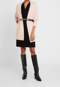 Eine Frau trägt ein schwarzes Kleid mit einem flauschigen, hellrosa Cardigan, der auf Taillenhöhe gebunden ist, und kniehohen schwarzen Lederstiefeln. Sie steht auf einem weißen Boden.