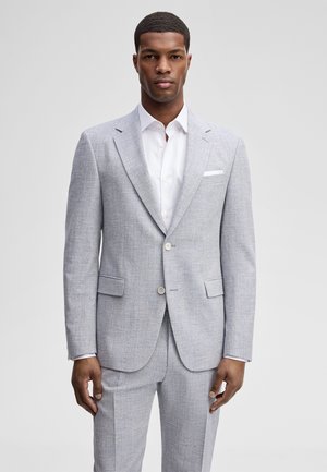 Man die een lichtgrijs pak draagt met een inkeping op het revers, een wit overhemd en een pochet, staand tegen een effen grijze achtergrond.