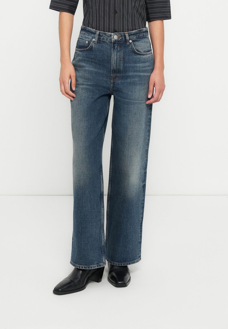 Femme portant un jean bleu délavé à jambes droites, des bottes noires à talons, et une chemise rayée noire et blanche rentrée, debout devant un fond blanc.