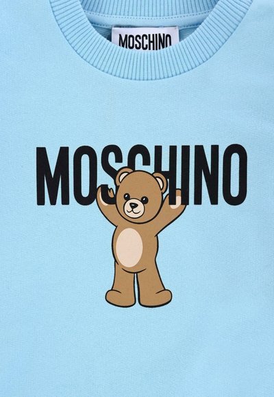 Felpa azzurra con testo nero "MOSCHINO" e grafica di un orsetto dei cartoni animati. L'orsetto ha un colore marrone chiaro con una pancia e braccia crema alzate.