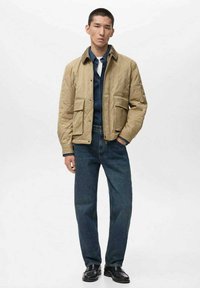 Giacca trapuntata beige con due tasche frontali, indossata sopra una camicia di jeans, abbinata a jeans blu e scarpe nere. Tessuto strutturato, design casual.