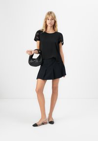 Donna bionda in piedi, che indossa una maglietta nera con maniche a occhiello, una gonna plissettata nera e scarpe slingback bicolore, mentre tiene una piccola borsa nera su sfondo bianco.