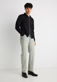 Νεαρός άνδρας στέκεται με το χέρι στην τσέπη φορώντας μαύρο πουκάμισο με φερμουάρ, ανοιχτοχρωμα τζιν και μαύρα δερμάτινα μποτάκια, με φόντο λευκό.