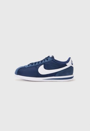 Marineblå Nike sneaker med hvid swoosh-logo, hvid sål med blå stribe, snøredesign og tekstureret tåhætte set fra siden.