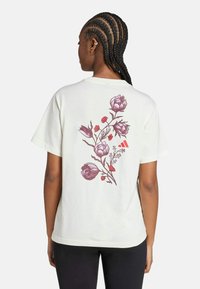 Camiseta de algodón blanca con una ilustración floral en púrpura y rojo en la parte posterior. Diseño de mangas cortas y corte relajado.