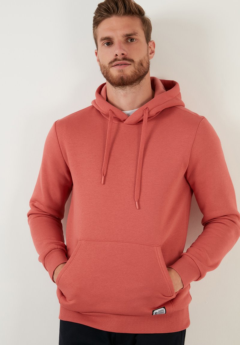Sudadera color coral hecha de tela suave, con un bolsillo frontal, capucha con cordones, puños acanalados y un pequeño parche con logo en la parte inferior.