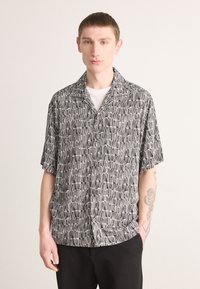 Black and white short-sleeve shirt featuring a geometric pattern, with a relaxed fit and collar. Styled over a white undershirt.