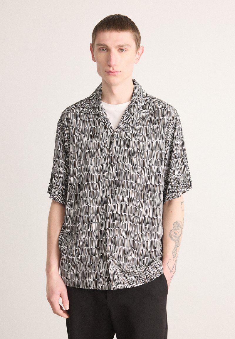 Black and white short-sleeve shirt featuring a geometric pattern, with a relaxed fit and collar. Styled over a white undershirt.
