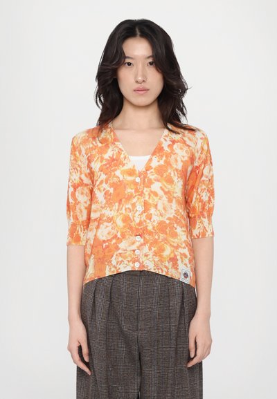 Cardigan floral orange avec un col en V, des manches courtes et une fermeture à boutons. Présente un ourlet côtelé et un tissu texturé. Associé à un pantalon à carreaux.