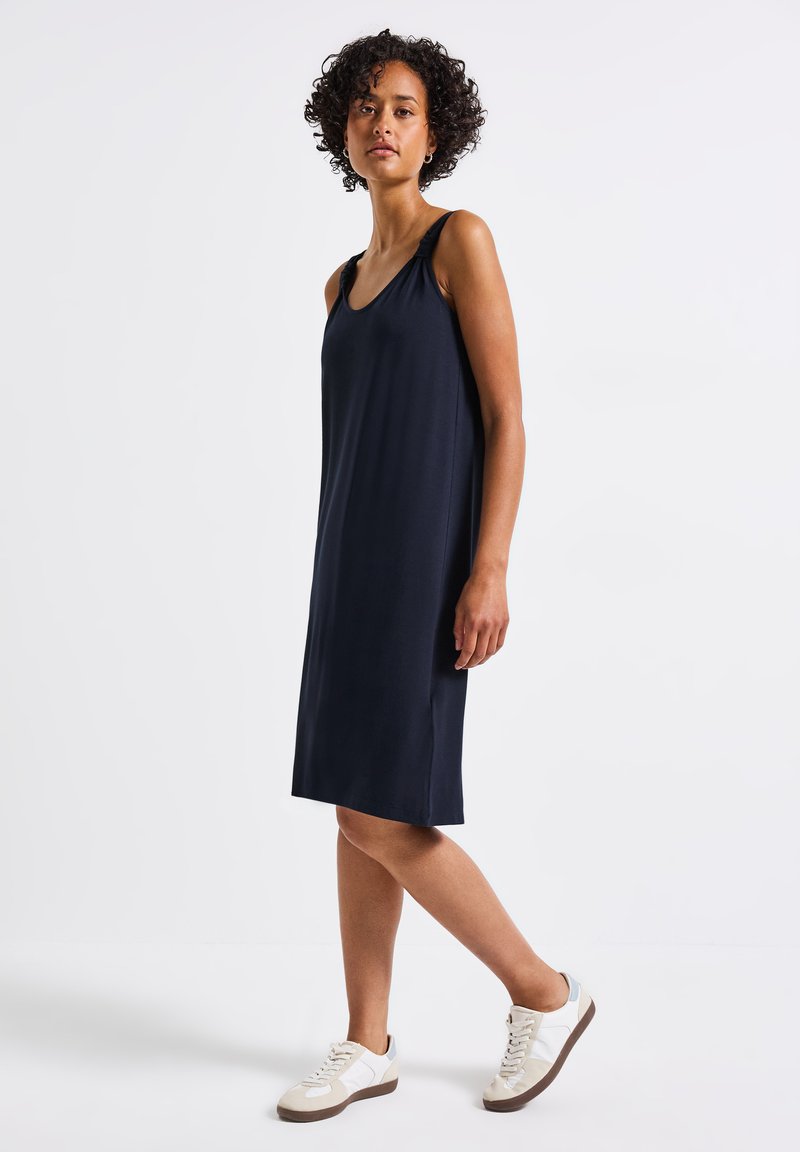Granatowa sukienka bez rękawów z marszczonymi detalami na ramionach, wykonana z gładkiego materiału. Połączona z jasnymi trampkami dla uzyskania swobodnego wyglądu.