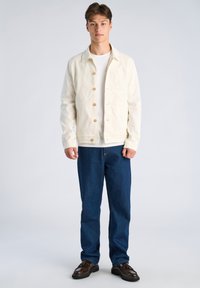 Beżowa kurtka dżinsowa z zapięciem na guziki i pojedynczą kieszenią na piersi, noszona na białej koszulce, zestawiona z ciemnoniebieskimi dżinsami i brązowymi butami.
