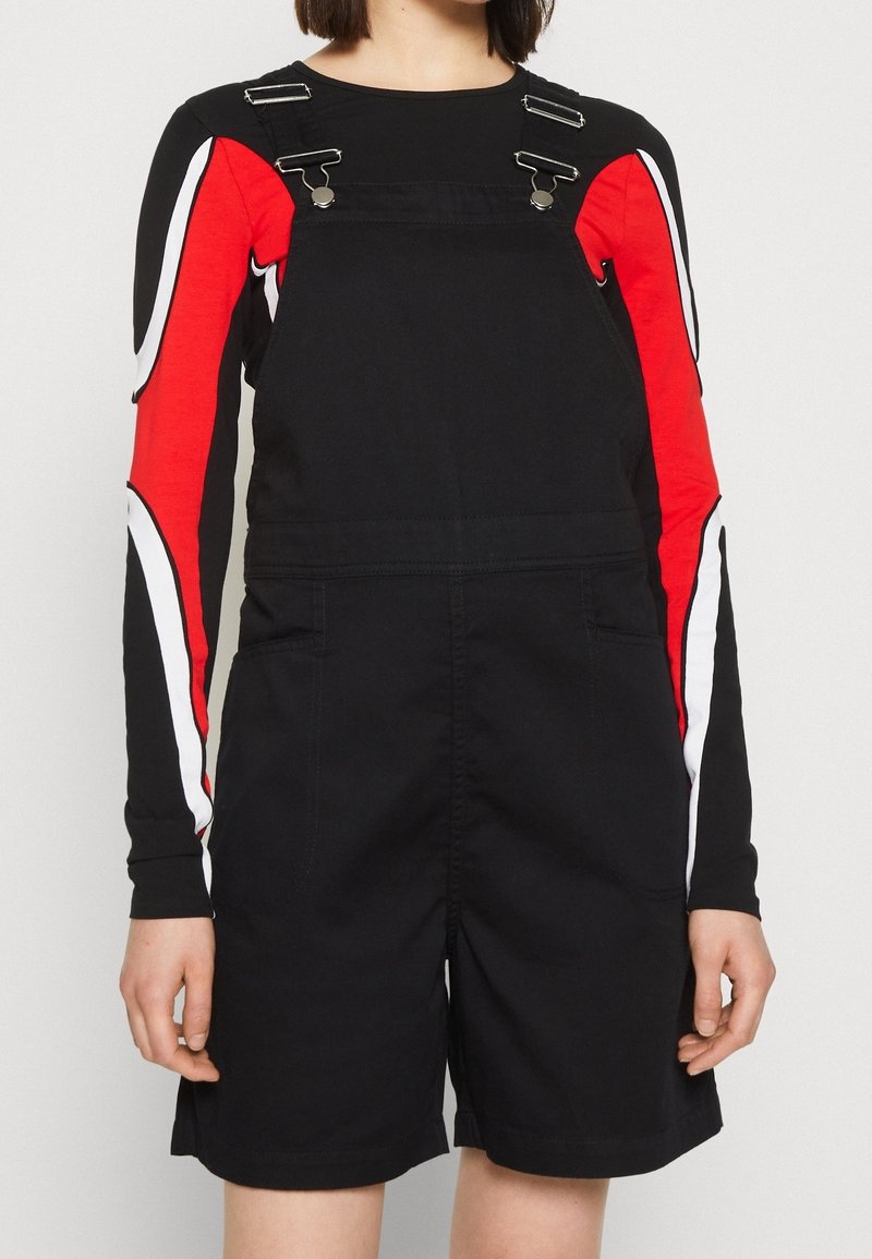 Svart overallklänning med justerbar silverdetalj. Långärmad skjorta med röda och vita böjda mönster på ärmarna. Material i bomullsblandning.