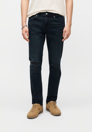 Persoon met zwarte gescheurde skinny jeans, lichtbeige shirt, zilveren armband en beige veterkuiten op een witte achtergrond.
