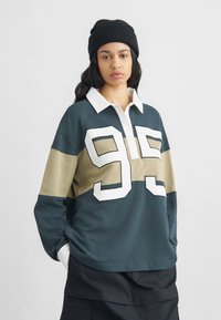 Μπλούζα πόλο με μακριά μανίκια σε σκούρο πράσινο με μπεζ και λευκές οριζόντιες ρίγες. Διαθέτει μεγάλο λευκό αριθμό "95" στο μπροστινό μέρος και λευκό γιακά.