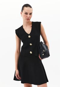 Schwarzes ärmelloses Oberteil mit V-Ausschnitt und drei goldenen Knöpfen, kombiniert mit einem ausgestellten schwarzen Rock. Das Model hält eine schwarze Handtasche.