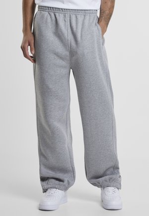 Homme portant un pantalon de survêtement gris ample avec des poignets élastiques, des baskets blanches et une chemise blanche, debout avec une main dans la poche.