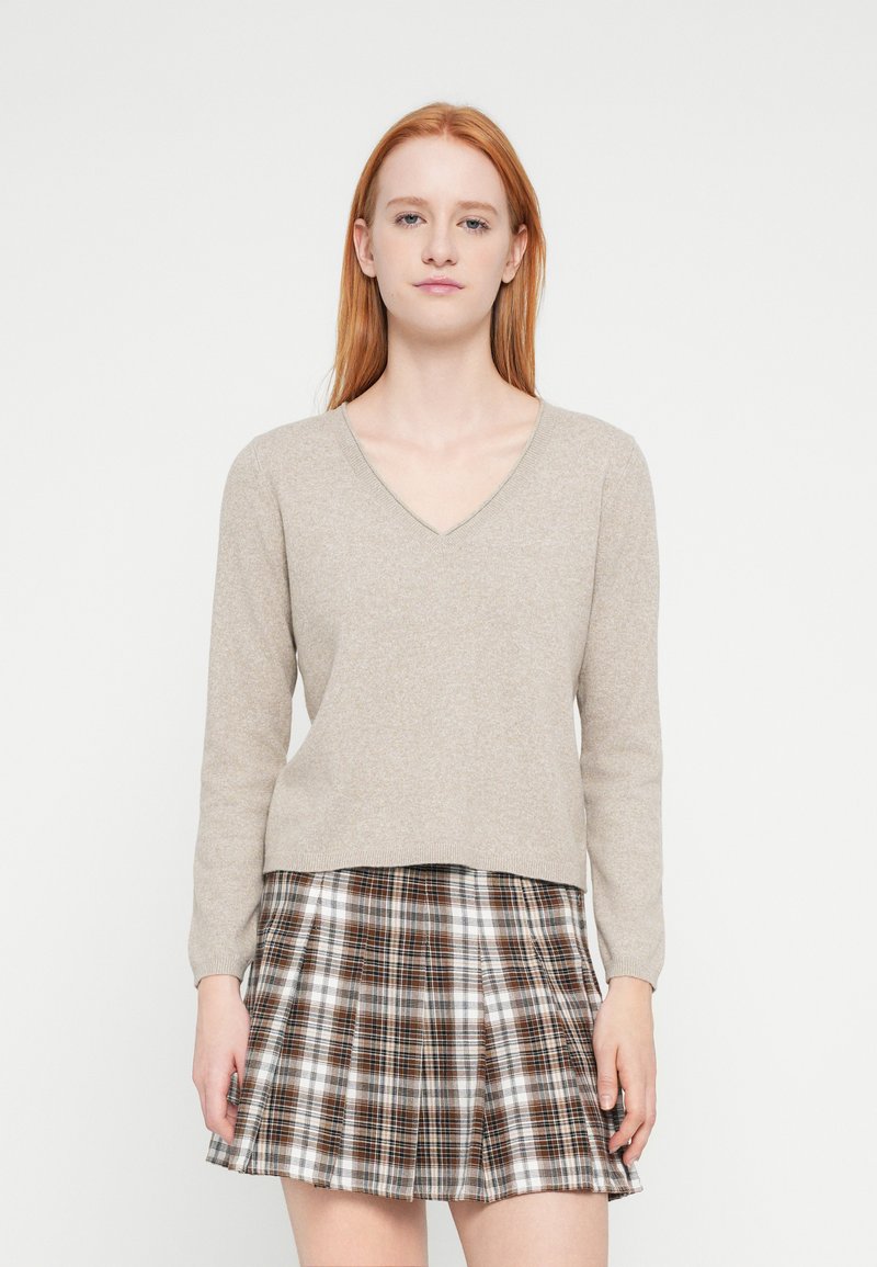 Pull beige à col en V avec manches longues, associé à une mini-jupe plissée à carreaux dans des tons marron, blanc et noir. Texture de tissu lisse.