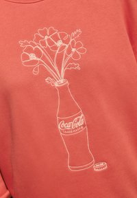 Korallenfarbener Sweatshirt mit weißer gestickter Darstellung einer Coca-Cola-Flasche, umgeben von Blumen und einem Detail des Flaschenverschlusses. Weiche Textur und lässiger Schnitt.