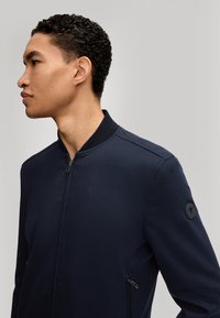 Navy bomberjack met een ritssluiting aan de voorkant, geribbelde kraag en twee zijzakken met rits. Bevat een ronde logopatch op de linkermouw. Glad textuur.