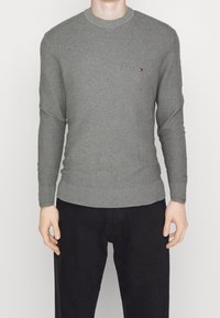 Pull gris en tricot à manches longues, col rond, avec poignets et ourlet côtelés. Petit logo sur la poitrine. Associé à un pantalon noir.