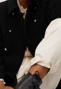 Abrigo negro cruzado con botones plateados, con una textura suave. Combinado con un top de punto beige y pantalones blancos, resaltando un conjunto en capas.