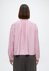 Rosa, lett bluse med en teksturert overflate, løs passform, lange ermer og detalj med samlet bak. Kombinert med grå bukser.