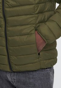 Main portant un gant brun placée dans la poche d'une veste matelassée vert olive avec une fermeture éclair noire, associée à un jean gris foncé.