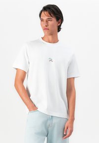 Jovem vestido com uma t-shirt branca de manga curta com um pequeno gráfico colorido no peito e jeans de ganga azul claro.