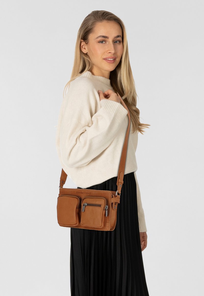 Borsa a tracolla in pelle marrone con due tasche frontali, chiusura con zip, tracolla regolabile e una texture liscia. Indossata con un maglione color crema.