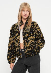 Joven mujer vestida con una chaqueta de forro polar con estampado floral en negro y marrón claro, un top corto blanco y pantalones negros, de pie con una mano en el bolsillo.