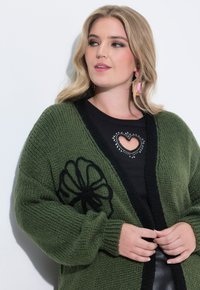 Grüne Strickjacke mit schwarzem Blumenmuster, offene Vorderseite und gerippte Bündchen, kombiniert mit einem schwarzen Top mit Herz-Ausschnitt und Verzierungen.