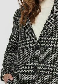 Kabát s vzorem houndstooth v černé a šedé barvě, s klopami, dvěma černými knoflíky a měkkou texturou. Pod ním bílý rolák.
