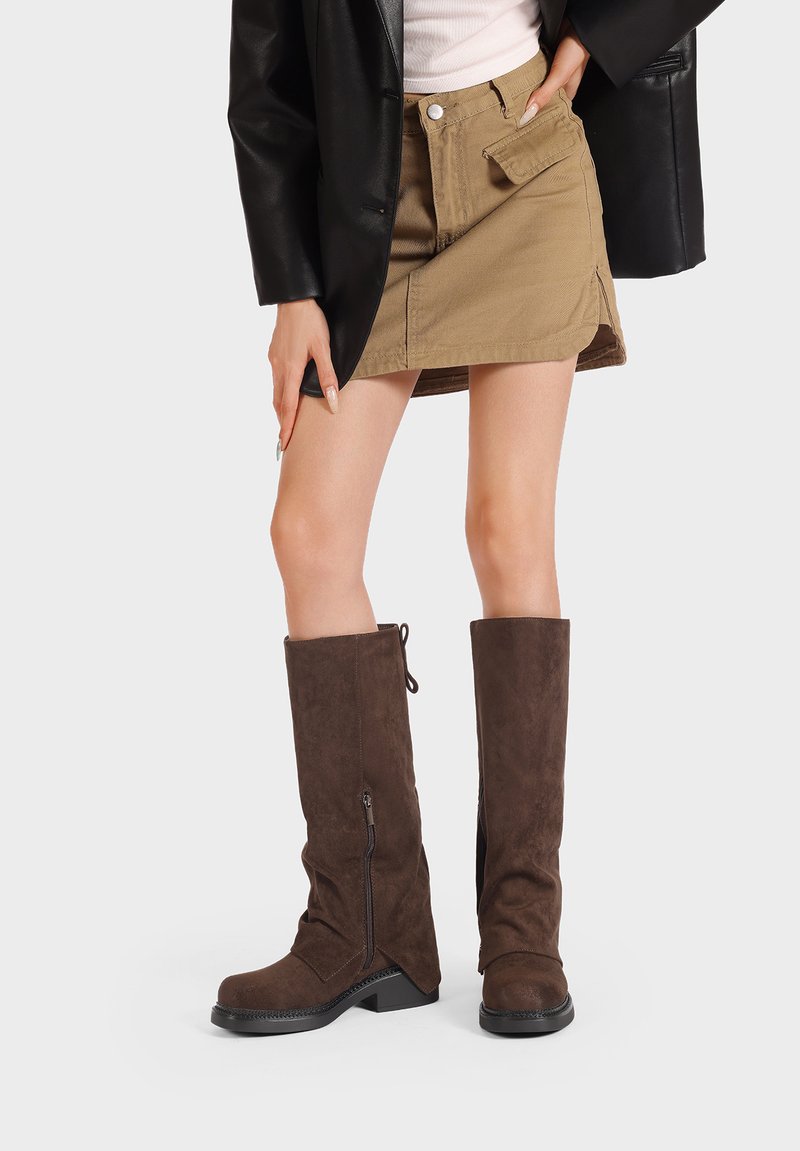 Stivali alti in suede marrone, con cerniera laterale, punta tondeggiante, tacco a blocco e dettagli in superficie texturizzati. Abbinati a una giacca nera e una gonna khaki.