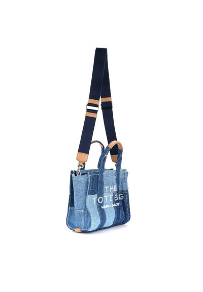 Denim tote bag with navy shoulder strap, featuring varying blue tones, beige leather accents, and white text reading "THE TOTE BAG."
