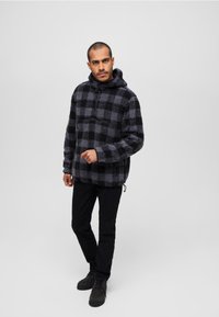 Fleece pulover s kapuco v črno-sivem karo vzorcu z polovičnim zadrgo, sprednjim žepom in vrvico. V kombinaciji s črnimi hlačami in čevlji.