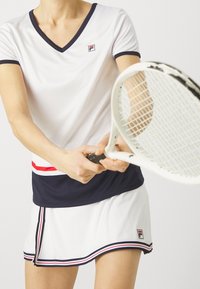 Bílý tenisový top s výstřihem do V, náměstíkovým lemem a logem Fila. V páru s bílou sukní s námořnickými a červenými akcenty. Raketou v ruce.