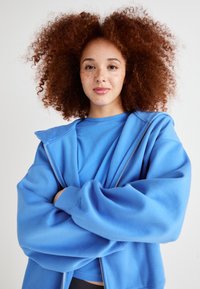 Blå zip-up hettegenser med en myk tekstur, lagd over en matchende T-skjorte. Modellen har krøllete rødt hår og armene krysset, noe som viser selvtillit.