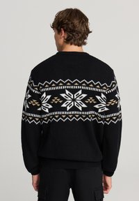 Czarny sweter wykonany na drutach z białym i złotym geometrycznym wzorem na klatce piersiowej, z prążkowanym kołnierzem i mankietami.