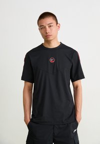 Melnā Nike T-krekla ar sarkanu sešstūru logotipu uz krūšu daļas, ar gludu tekstūru un īsām piedurknēm, apvienots ar melniem cargo šortiem.