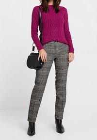 Frau trägt einen magentafarbenen, gestrickten Pullover, schwarze und weiße karierte Hose, schwarze Stiefeletten und trägt eine kleine schwarze Umhängetasche.