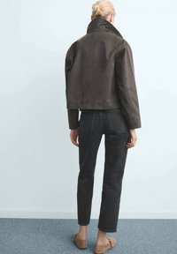 Mujer con cabello rubio recogido en un moño, vistiendo una chaqueta corta marrón oscura, jeans negros y zapatos planos beige, de pie contra una pared blanca.