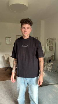 Camiseta negra de gran tamaño con texto blanco, combinada con jeans de color azul claro. El tono de piel es claro; las características incluyen tatuajes en los brazos.