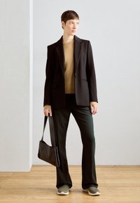 Blazer nero su misura, maglione marrone, pantaloni verde scuro e sneakers verde chiaro. Tiene una borsa nera. Tessuti morbidi, design strutturato.