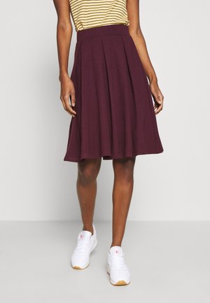 Personne portant une jupe plissée bordeaux à mi-longueur, un haut rayé jaune et des baskets blanches devant un fond clair.