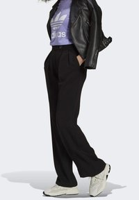 Svarta vida byxor med veckad framsida, kombinerat med en lila t-shirt med en vit logotyp och en svart läderjacka. Vita sportiga sneakers.