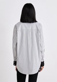 Chemise blanche à rayures verticales noires, à manches longues et à ourlet arrondi. Présente des poignets à boutons noirs et un design de col classique.