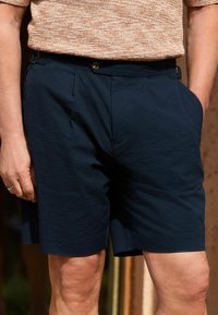 Shorts bleu marine en tissu texturé avec poches latérales et fermeture à bouton, présentant une coupe décontractée et un léger détail de pli.